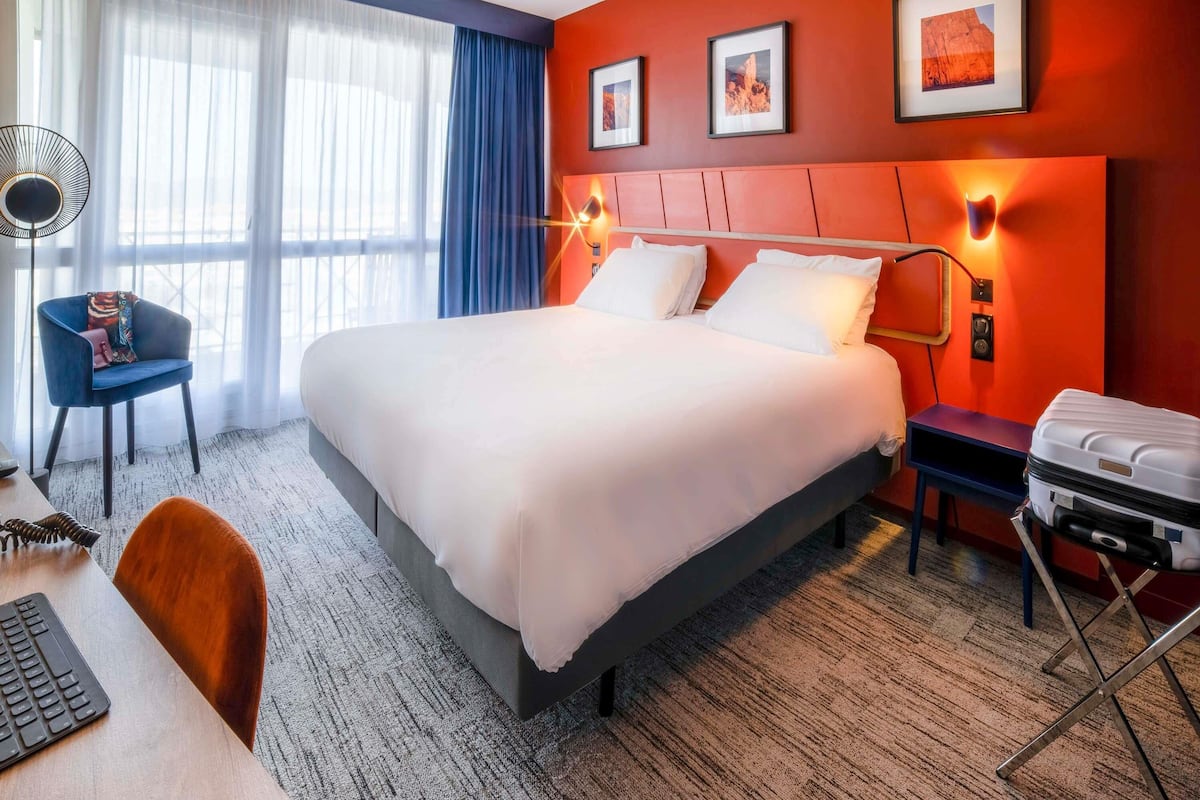 Ein Hotelzimmer mit einem großen Bett, einem Schreibtisch mit Tastatur, einem Sessel, einem Koffer und gerahmten Bildern an der Wand.