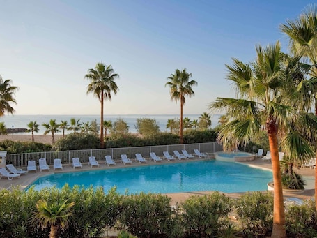 A pool area with lounge chairs and palm trees, overlooking a beach.. Mercure Thalasso and Spa Port Frejus