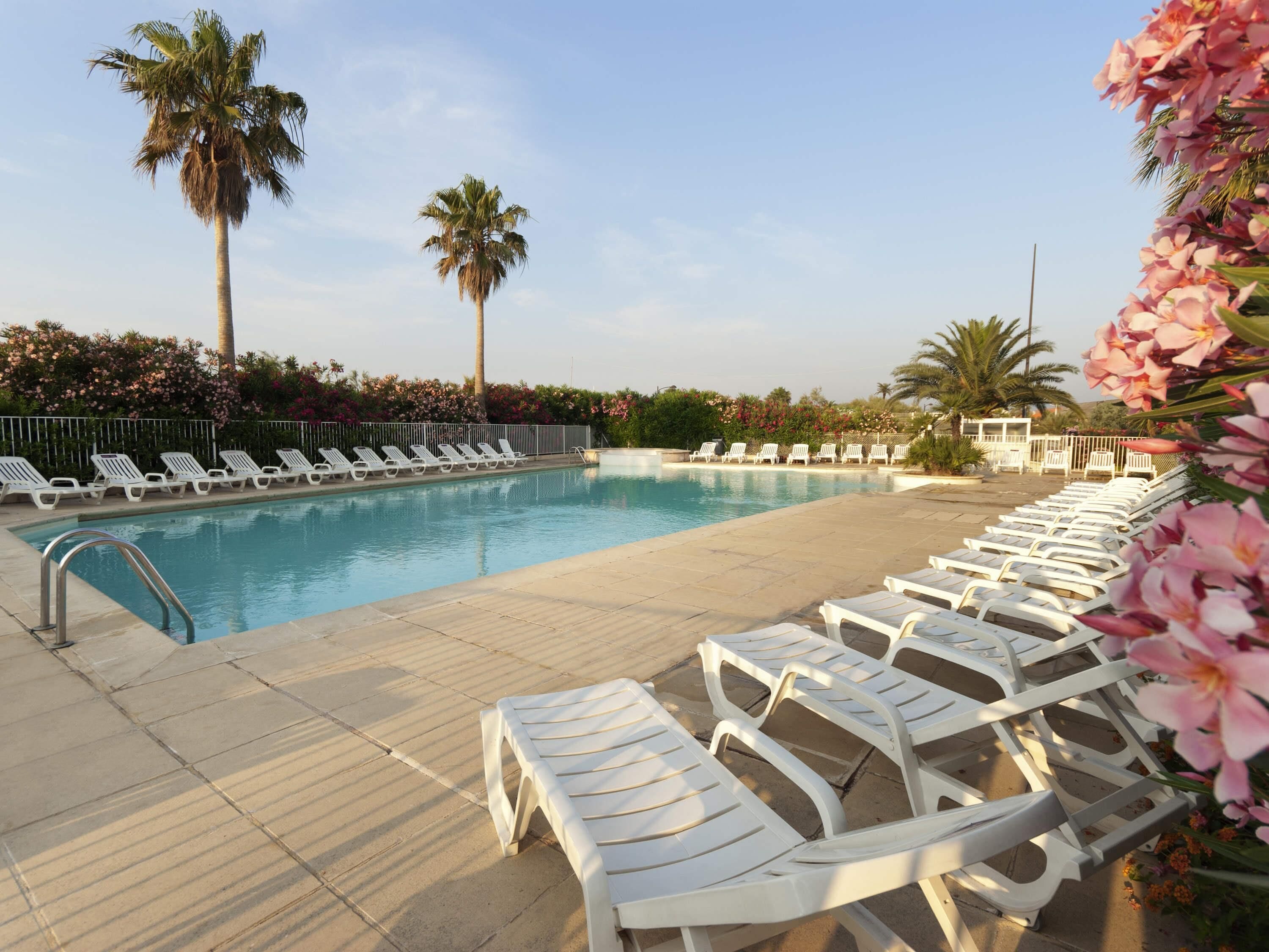 A pool with white lounge chairs, palm trees, and pink flowers.