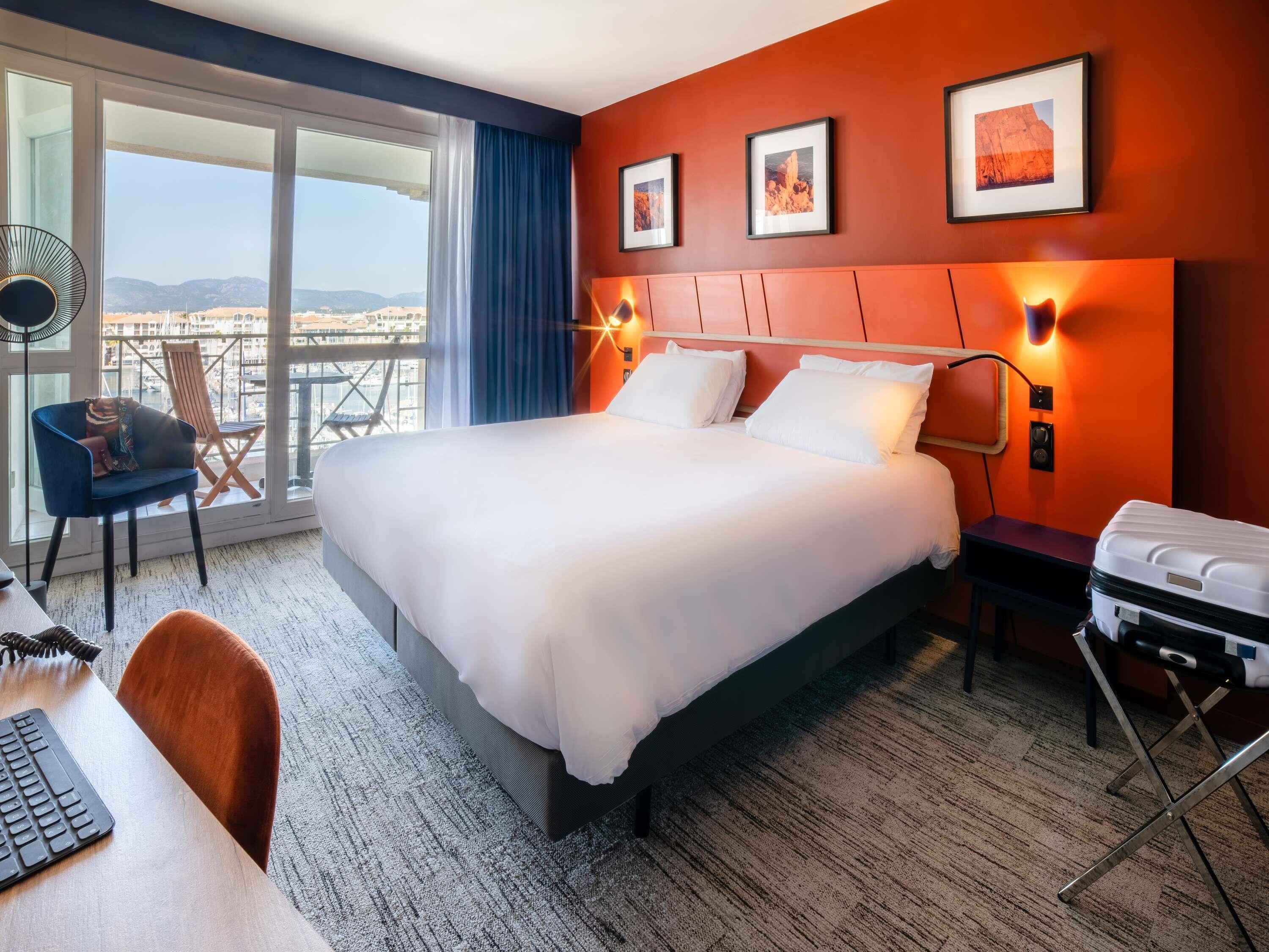 Ein Hotelzimmer mit einem großen Bett, einem Schreibtisch mit Tastatur, einem Stuhl, einem Koffer und einem Balkon mit Aussicht.