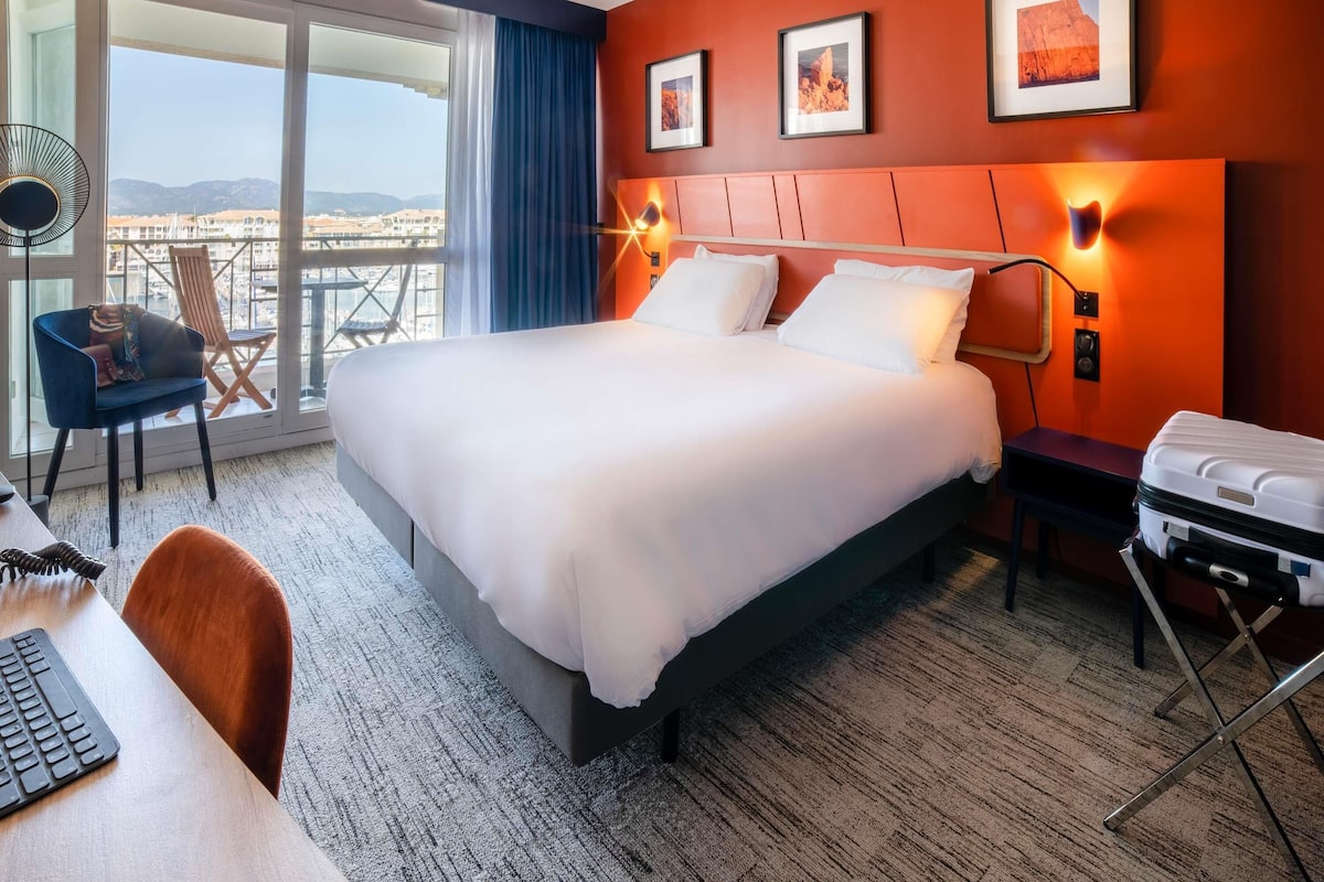 Ein Hotelzimmer mit einem großen Bett, einem Schreibtisch mit Tastatur, einem Stuhl, einem Koffer und einem Balkon mit Aussicht.
