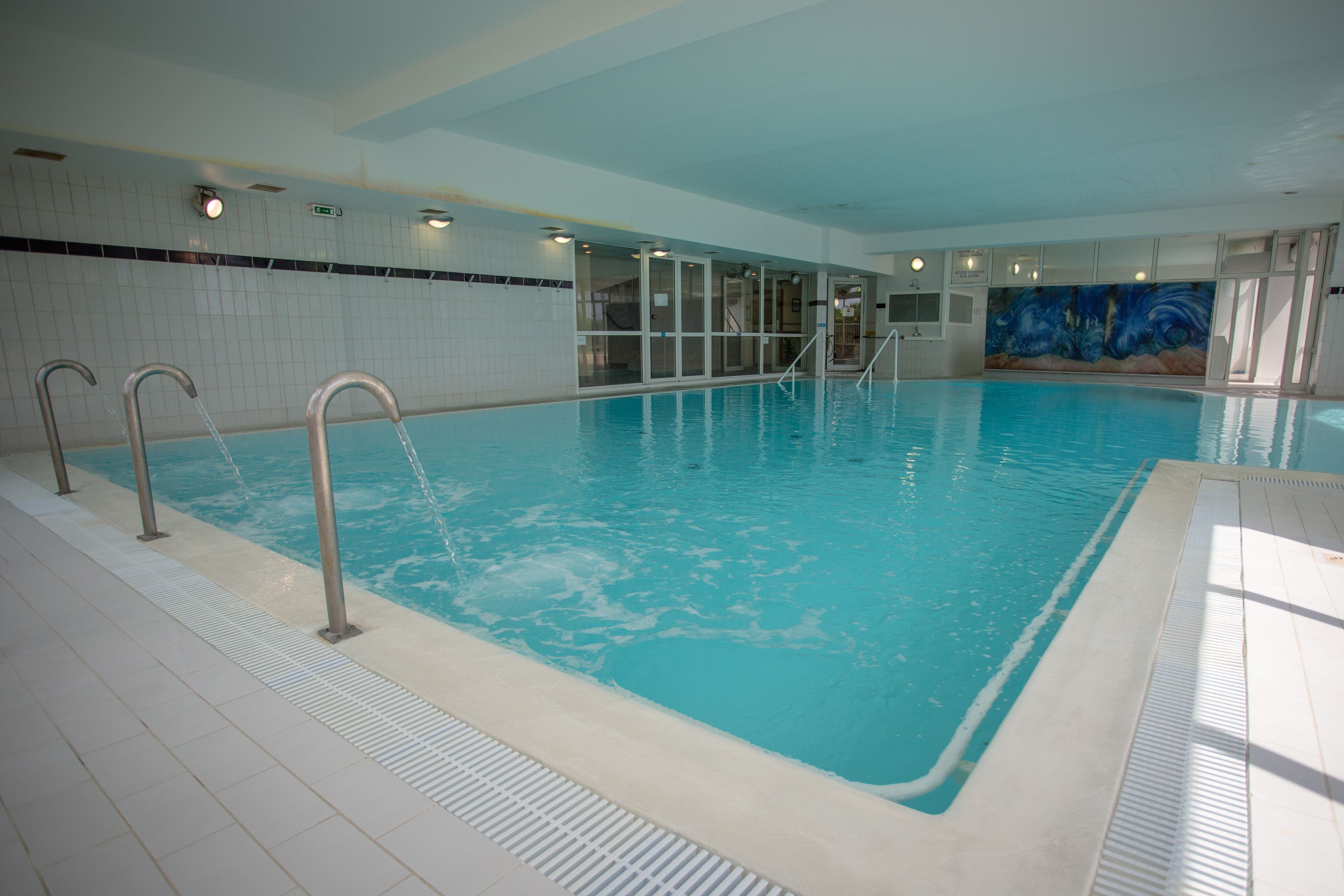Una piscina interior con agua cristalina, escaleras metálicas y suelo de baldosas.