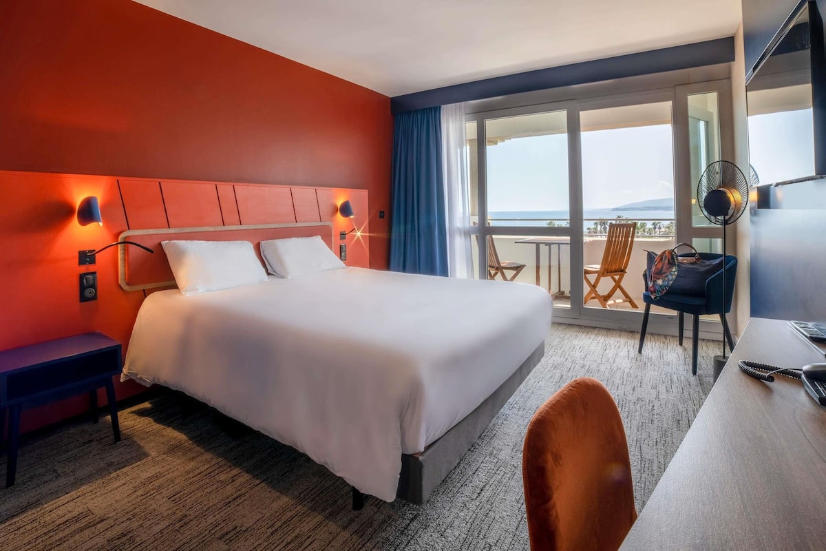 Ein Hotelzimmer mit Bett, Schreibtisch, Stuhl und einem Balkon mit Blick.