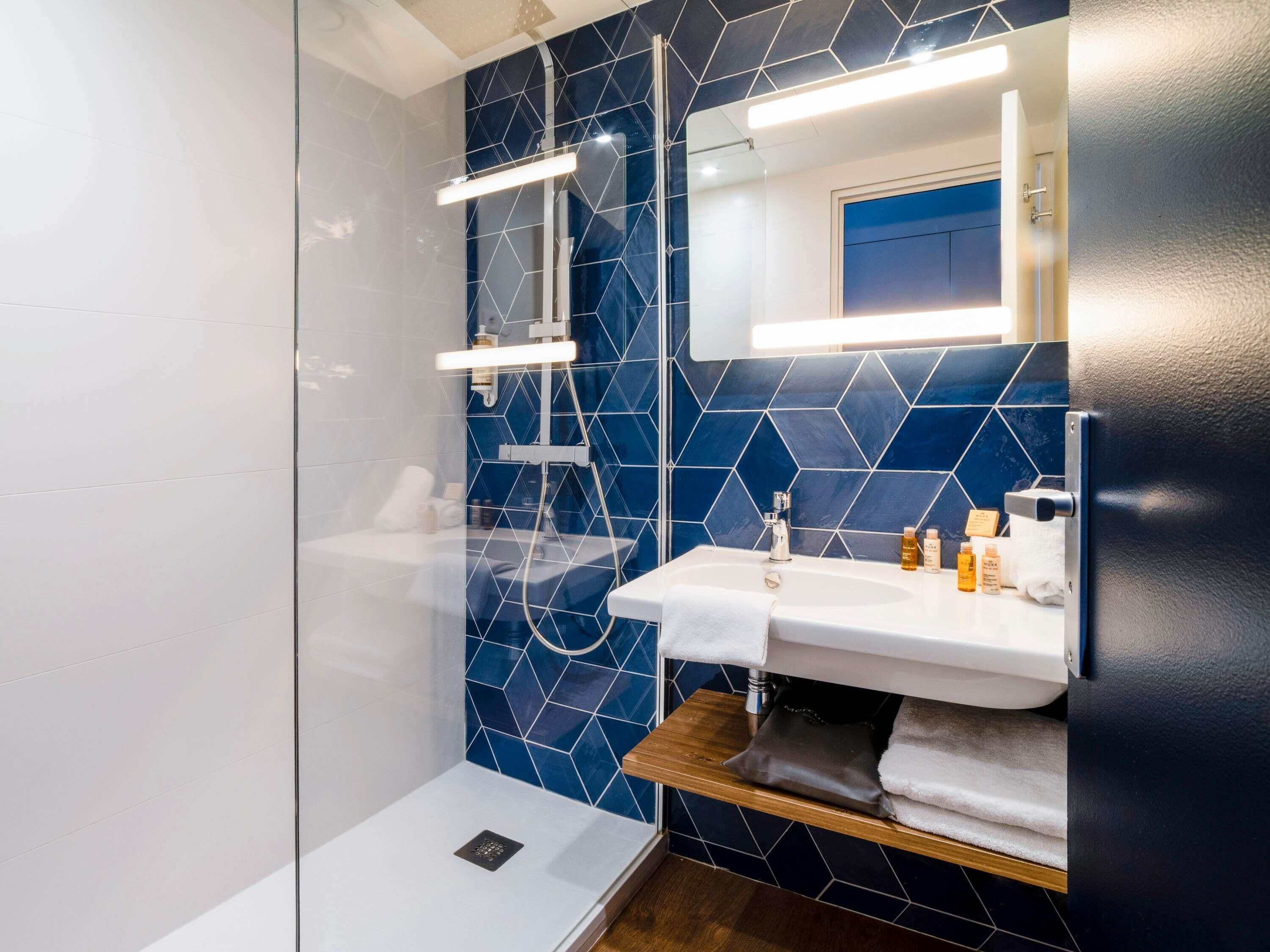 A modern bathroom with a glass shower enclosure, a white sink with a mirror above, and a blue-tiled wall.