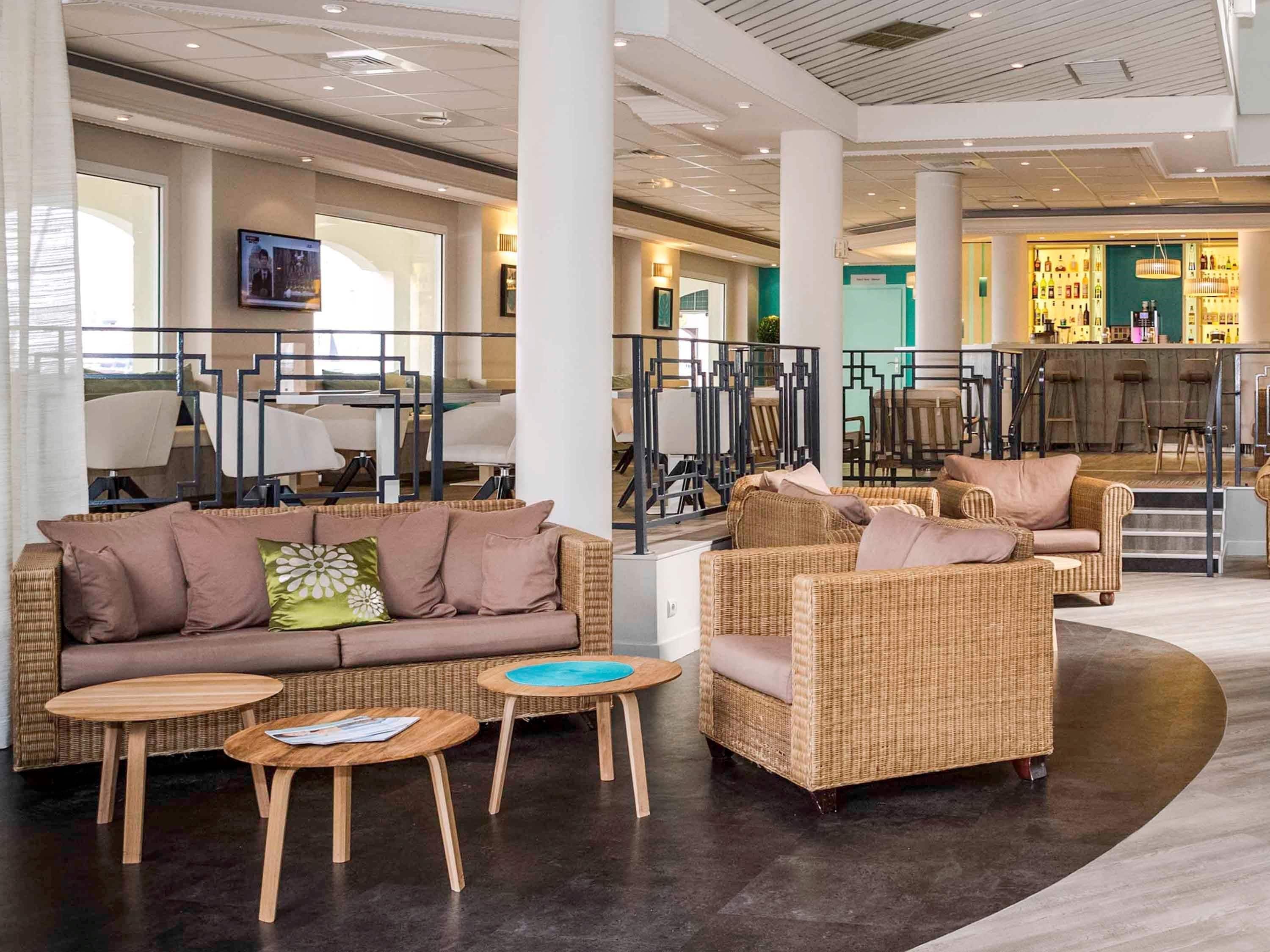 Un moderno vestíbulo de hotel con un área de estar que incluye un sofá y dos sillas, una zona de bar con taburetes y un televisor montado en la pared.