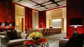 Un hall de hotel con alfombra roja, una recepción, un comedor con una mesa larga y sillas, y un área de descanso con sofás y una mesa baja.