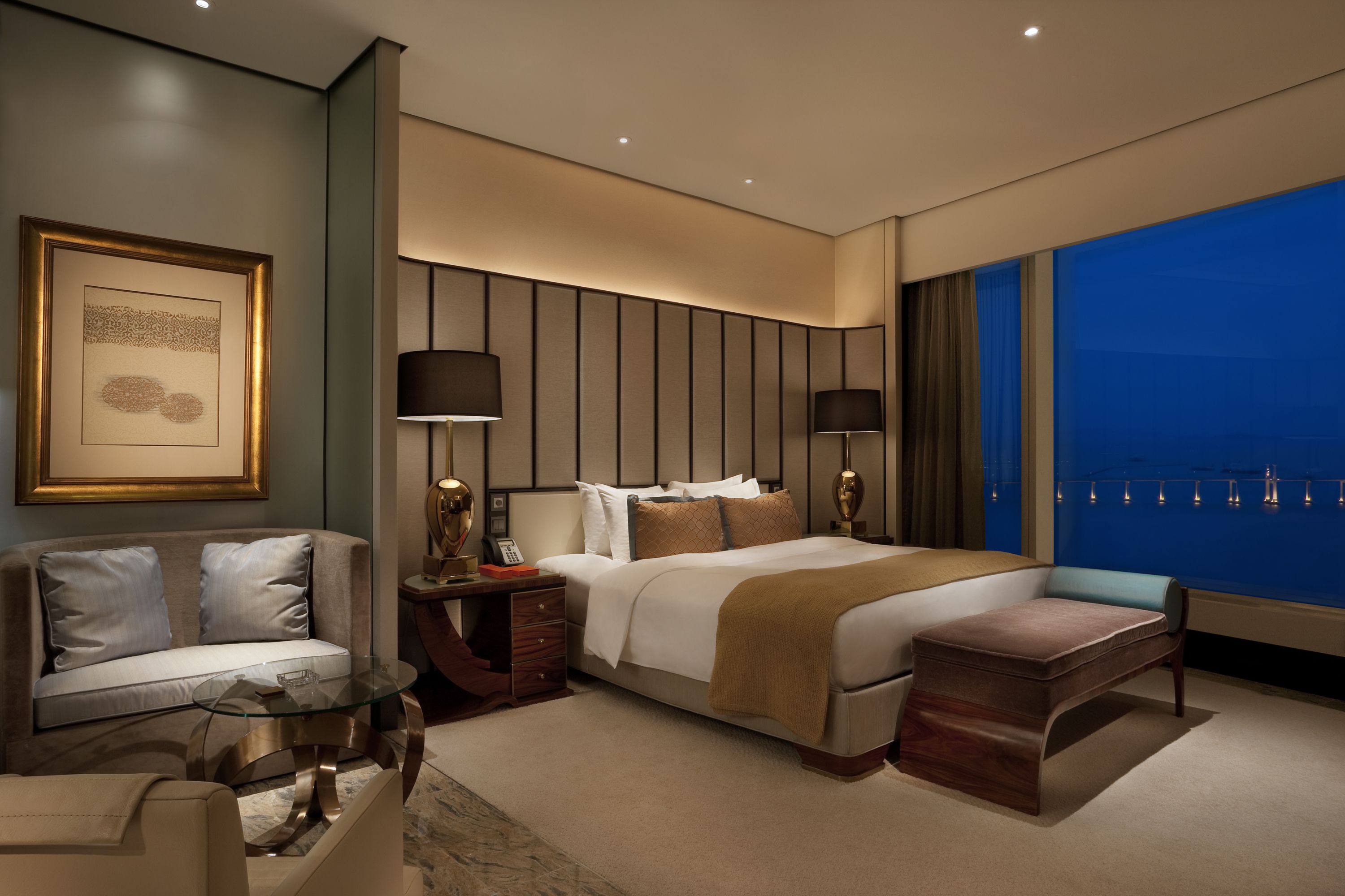 A modern hotel room with a large bed, a sofa, a glass coffee table, and a view of the city skyline.