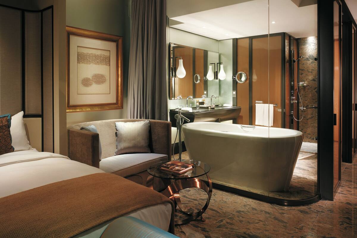 A modern hotel room with a large bed, a sitting area with a glass table, a bathtub, and a bathroom with a vanity and shower.