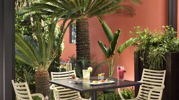 A table set with white chairs, a drink, and a plate of food in a lush outdoor setting with various plants and a palm tree.