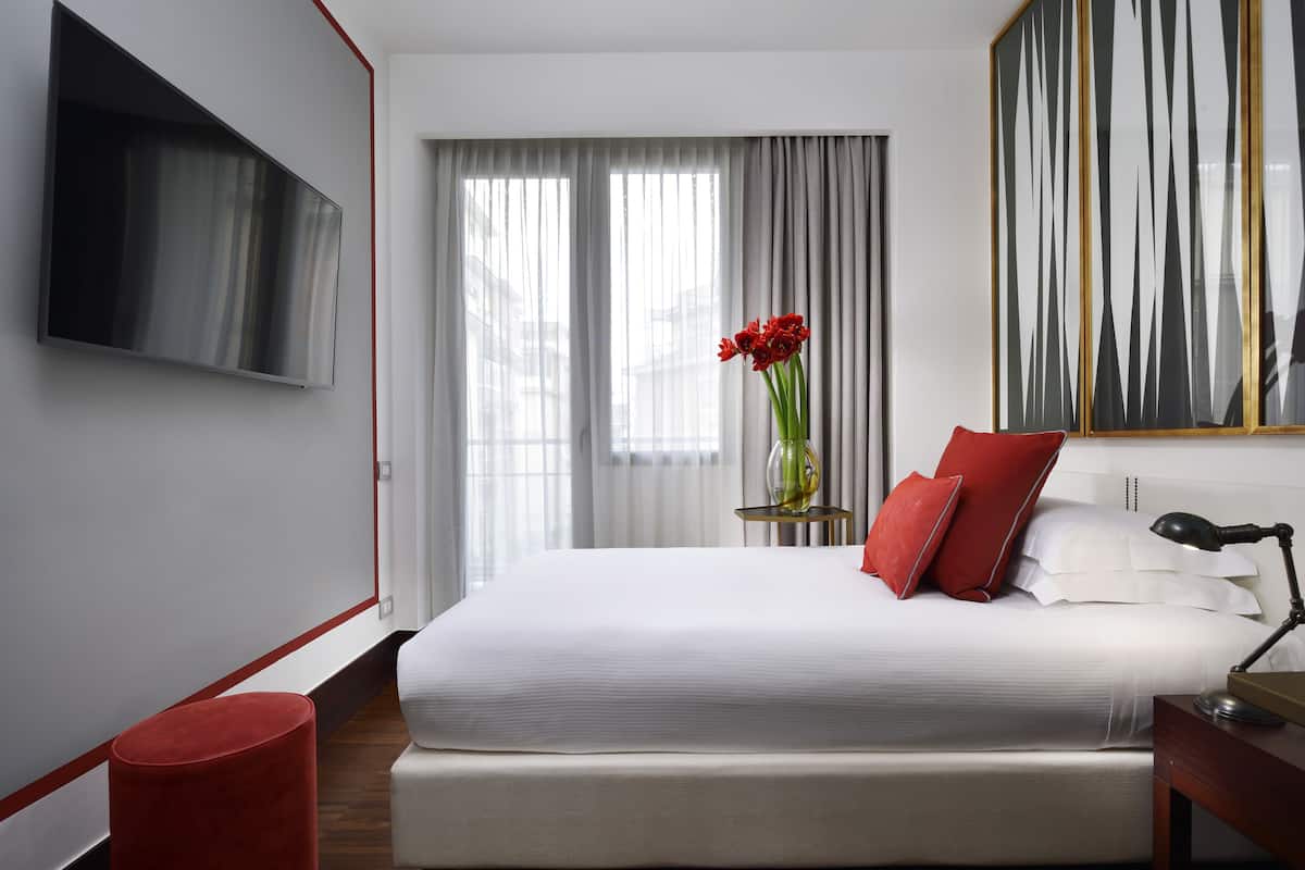A modern hotel room with a large bed, a flat-screen TV mounted on the wall, a vase with red flowers, and a large abstract painting on the wall.