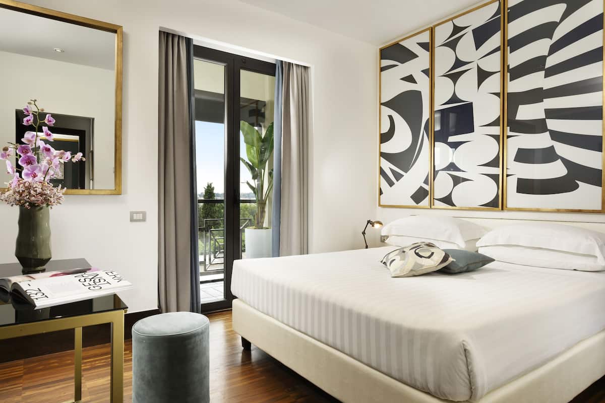 A modern hotel room with a large bed, a desk with a vase of flowers, a large mirror, and a view of the outdoors.