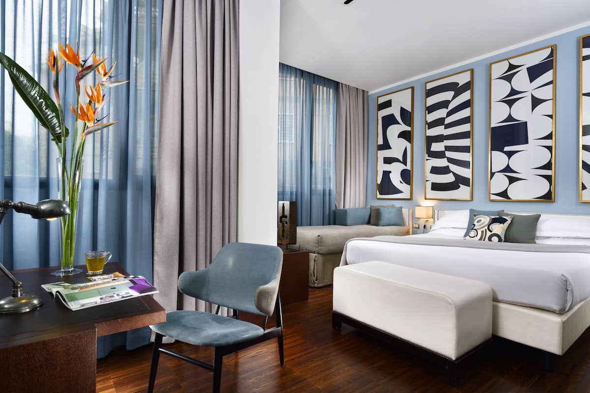 A modern hotel room with a bed, a desk with a lamp, a chair, and a large window with curtains.