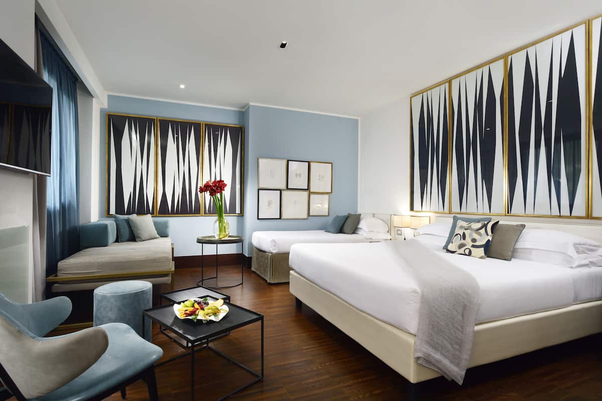 A hotel room with two beds, a sofa, a small table with fruit, and a vase of flowers.