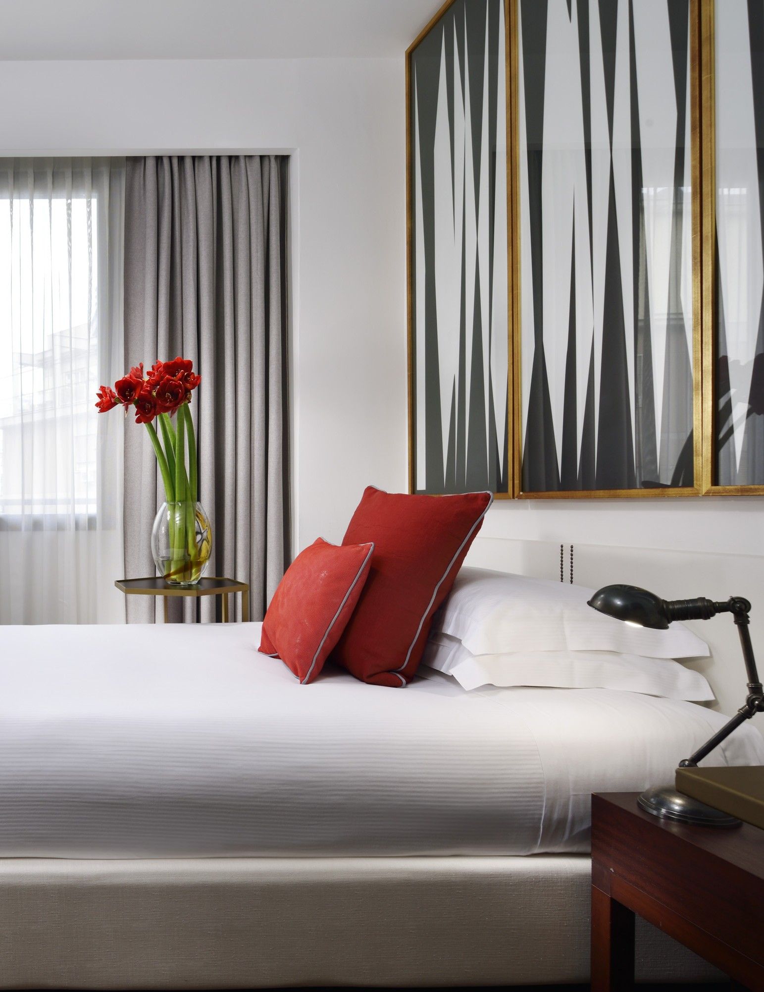 Una habitación de hotel con una cama, un jarrón de flores rojas, un cojín rojo, una mesita de noche con una lámpara y un cuadro abstracto grande en la pared.