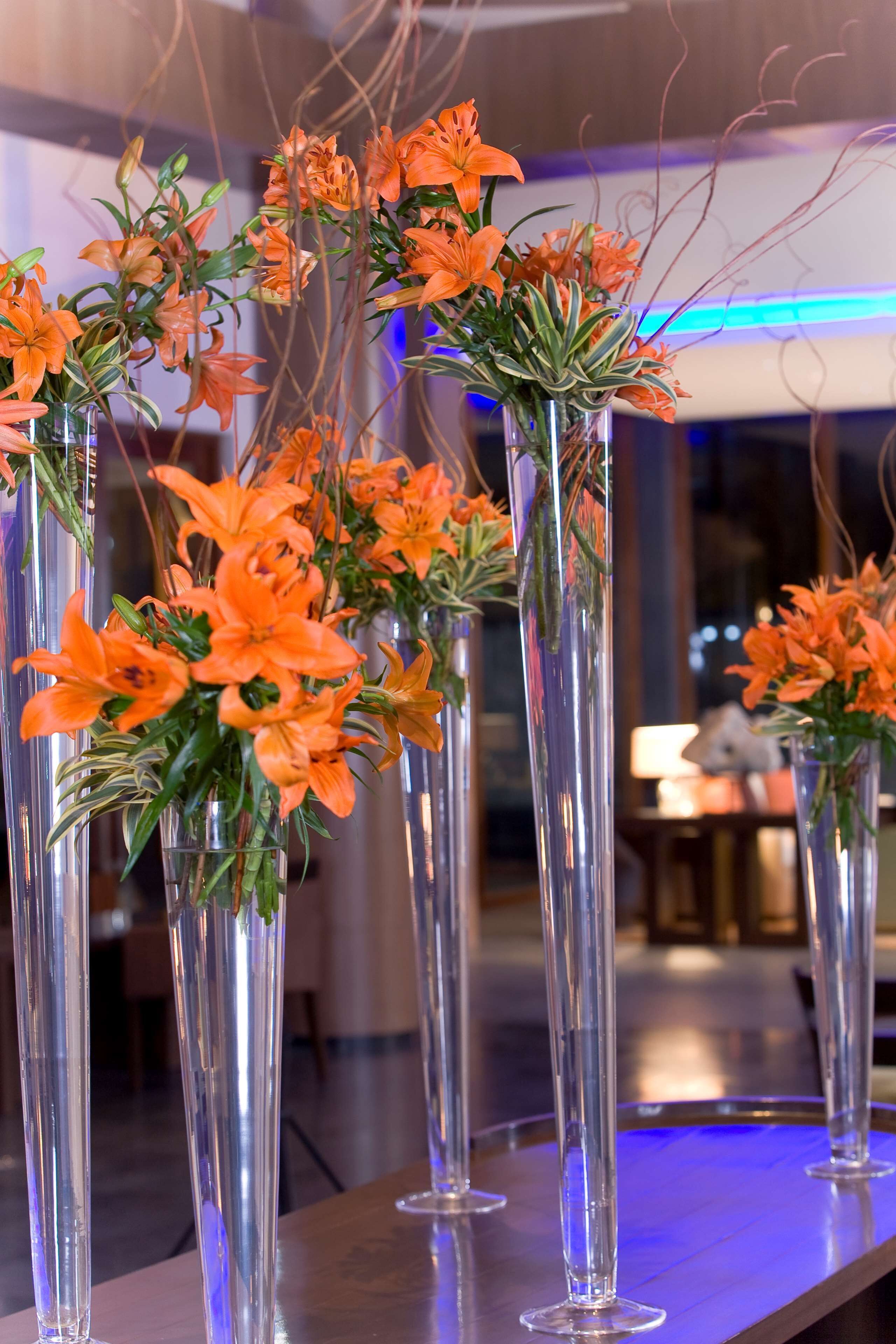 Tall, clear glass vases filled with orange flowers and green foliage, arranged on a reflective surface.