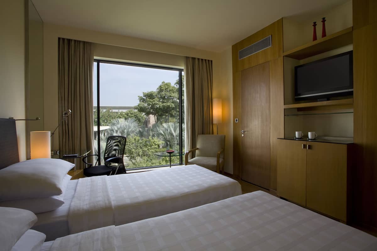 A hotel room with two beds, a TV, a desk with a lamp, and a view of greenery outside.