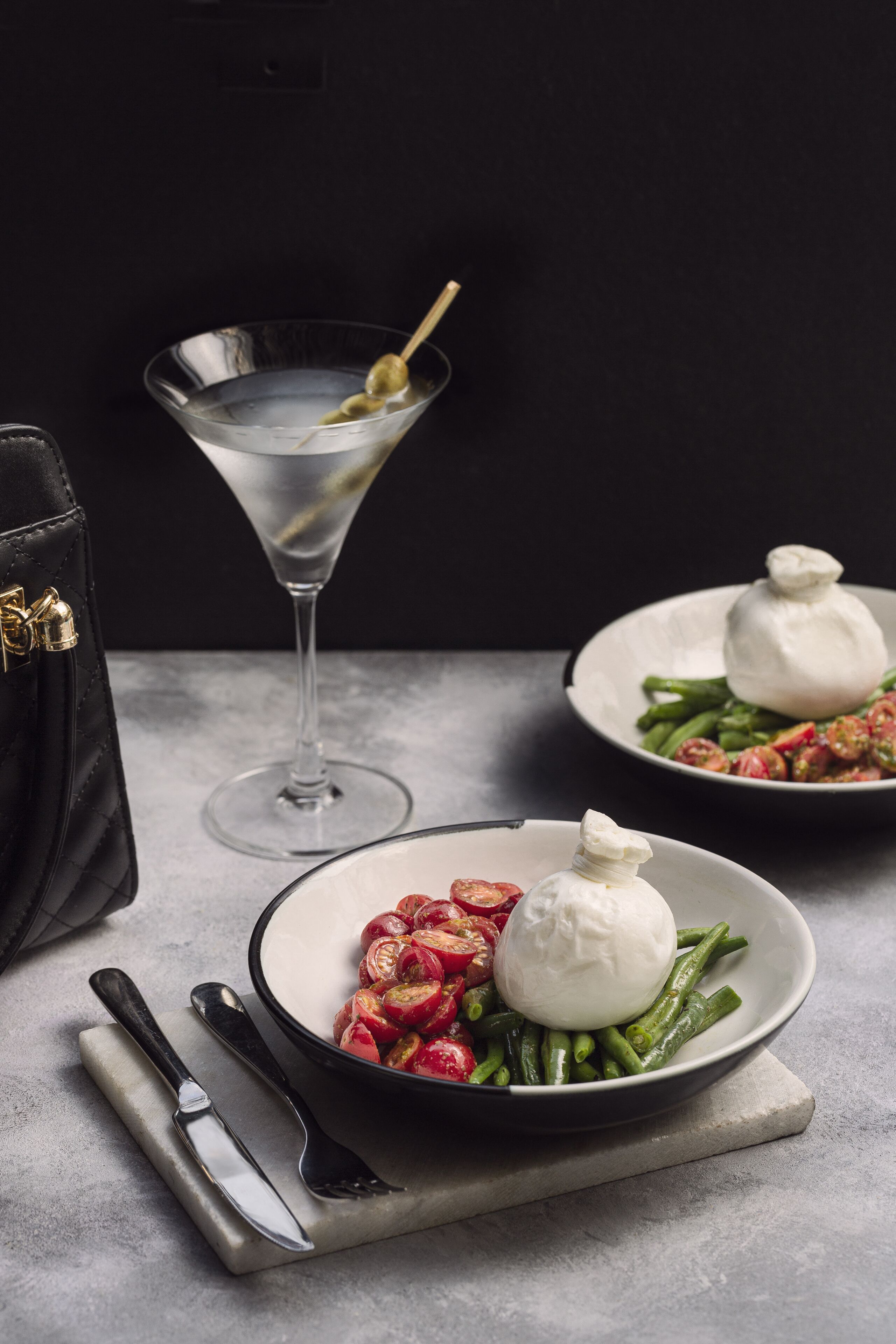 A martini glass with olives, a black handbag, and plates with green beans and cherry tomatoes.