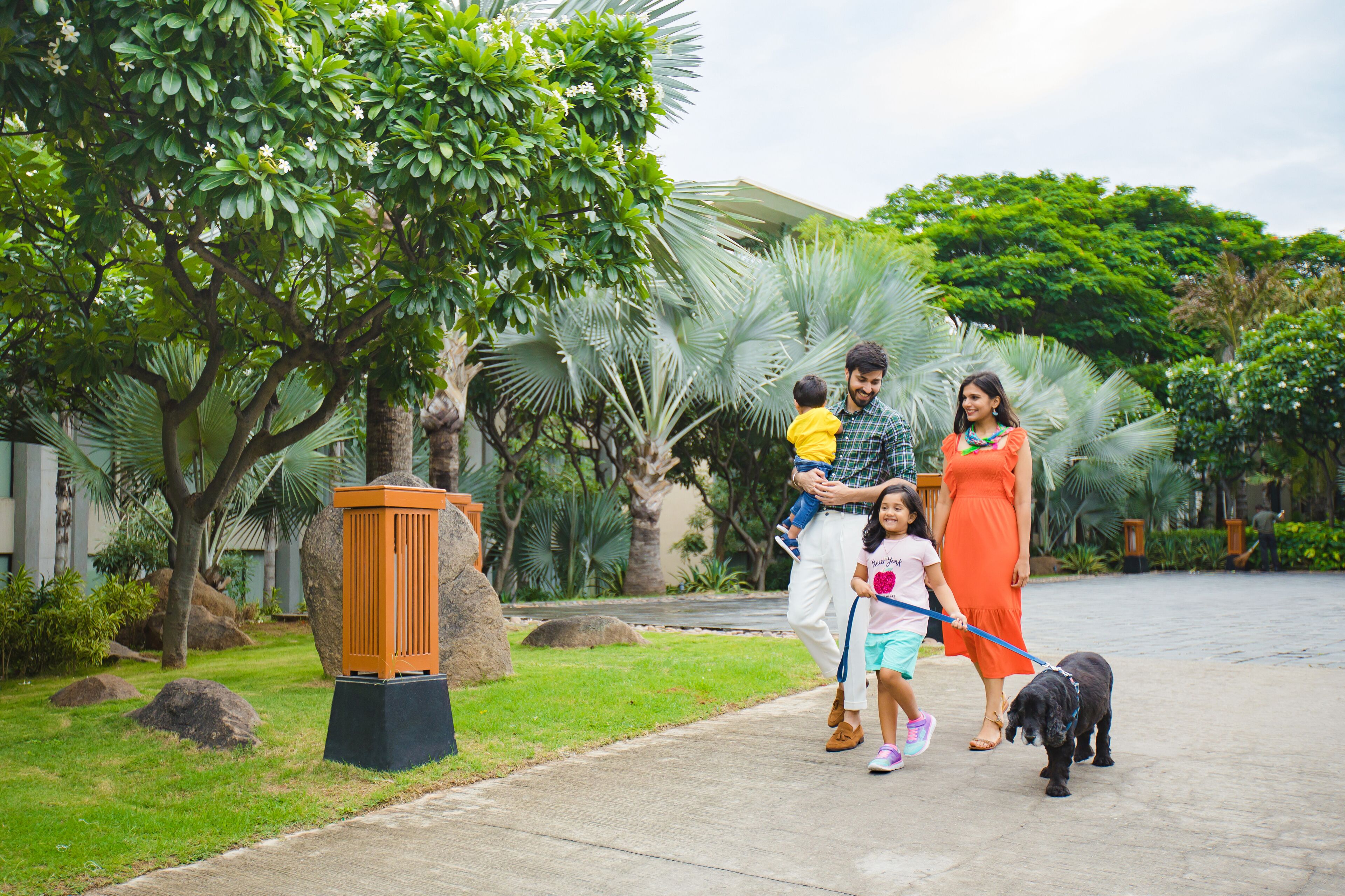 A family walking a dog on a paved path in a lush, green outdoor area with palm trees and a large planter.