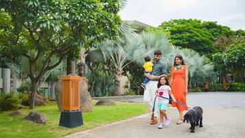 A family walking a dog on a paved path in a lush, green outdoor area with palm trees and a large planter.