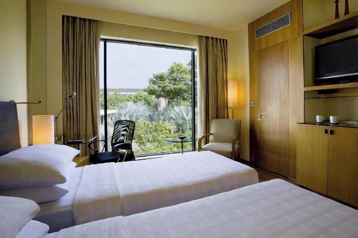 A hotel room with two beds, a TV, a chair, a small table, and a window with curtains.
