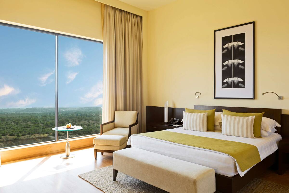 A hotel room with a large window, a bed with pillows, a chair, a small table, and a framed picture on the wall.