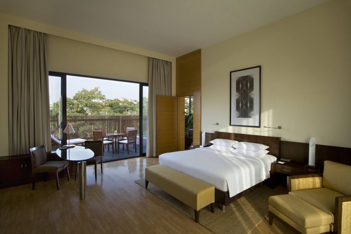 A hotel room with a large bed, a desk, a chair, a sofa, and a balcony with a view of trees.