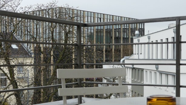 Un balcon avec une chaise blanche et une petite table, d’où l’on peut admirer un paysage urbain composé d’immeubles modernes et d’arbres sans feuilles.