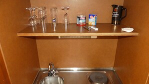 Un coin cuisine comprenant un évier, deux plaques de cuisson, une machine à café, une canette de bière et plusieurs verres disposés sur une étagère.