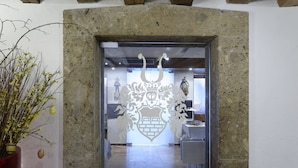 Une arche en pierre ornée d’un motif décoratif, un pot de fleurs et un aperçu d’une pièce où l’on aperçoit une table et des chaises.