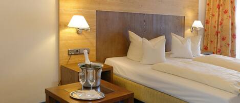 Habitación de hotel con cabecera de madera, dos lámparas de noche, cama con ropa blanca, una mesita de madera con un vaso y un plato, y alfombra azul estampada.