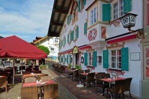 An outdoor café with wicker tables and chairs, a red umbrella, and traditional European architecture. - Hotel Alte Post (Oberammergau)