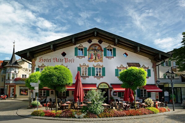 A traditional European building with a sign reading "Hotel Alte Post." It features a central entrance with a red awning, surrounded by outdoor seating and flower beds. - Hotel Alte Post (Oberammergau)