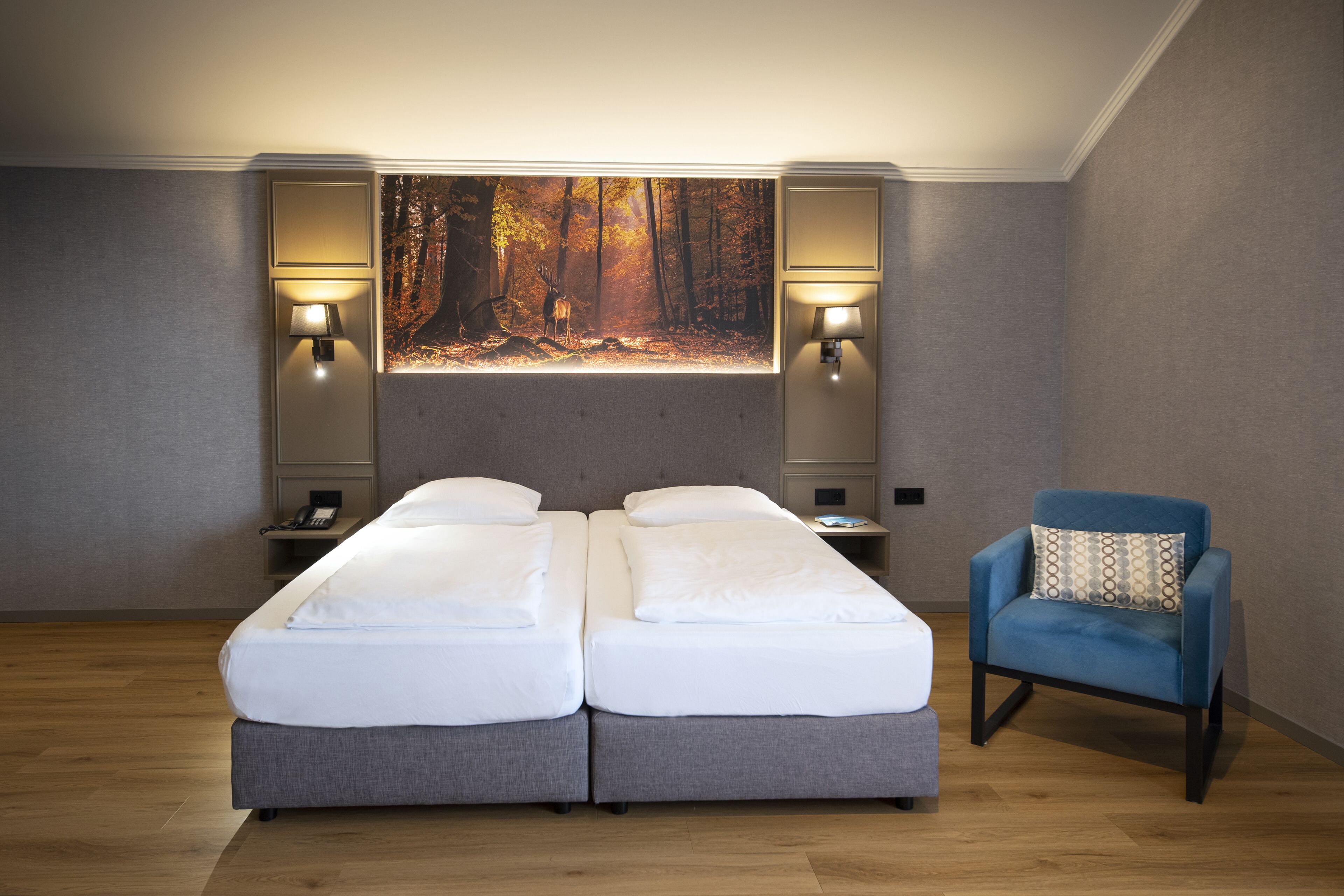 Un dormitorio con una cama doble, dos mesitas de noche, un revistero, un sillón azul y una obra de arte de gran tamaño con motivos forestales.