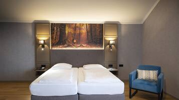 Un dormitorio con una cama doble, dos mesitas de noche, un revistero, un sillón azul y una obra de arte de gran tamaño con motivos forestales.