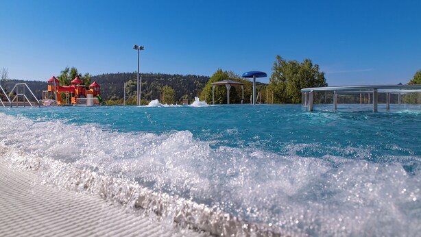 A swimming pool with water splashing, a sunshade, and a view of trees and hills in the background.