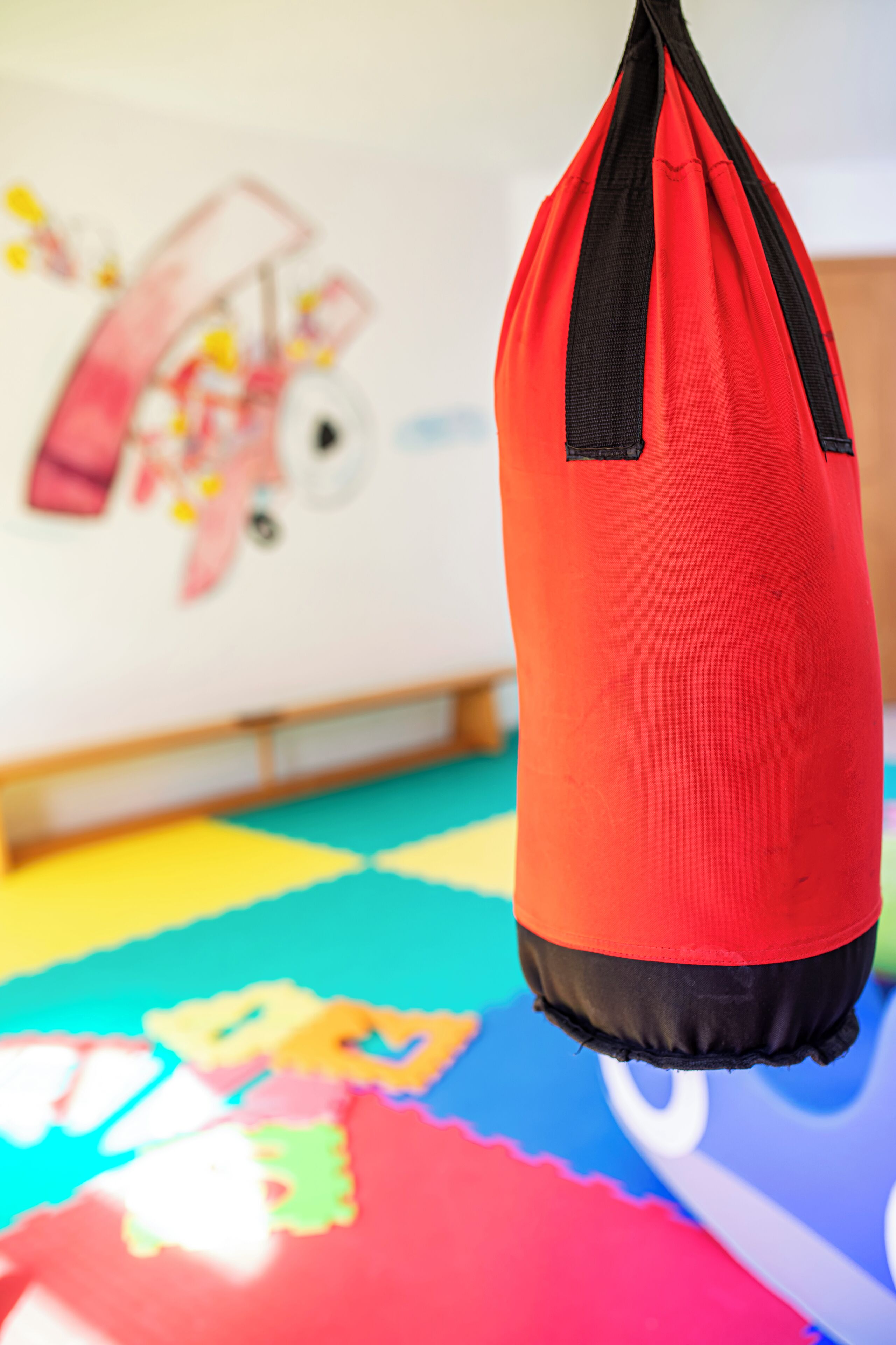 Un saco de boxeo rojo colgado en una habitación con esterillas de colores en el suelo y una pared con un personaje de caricatura.