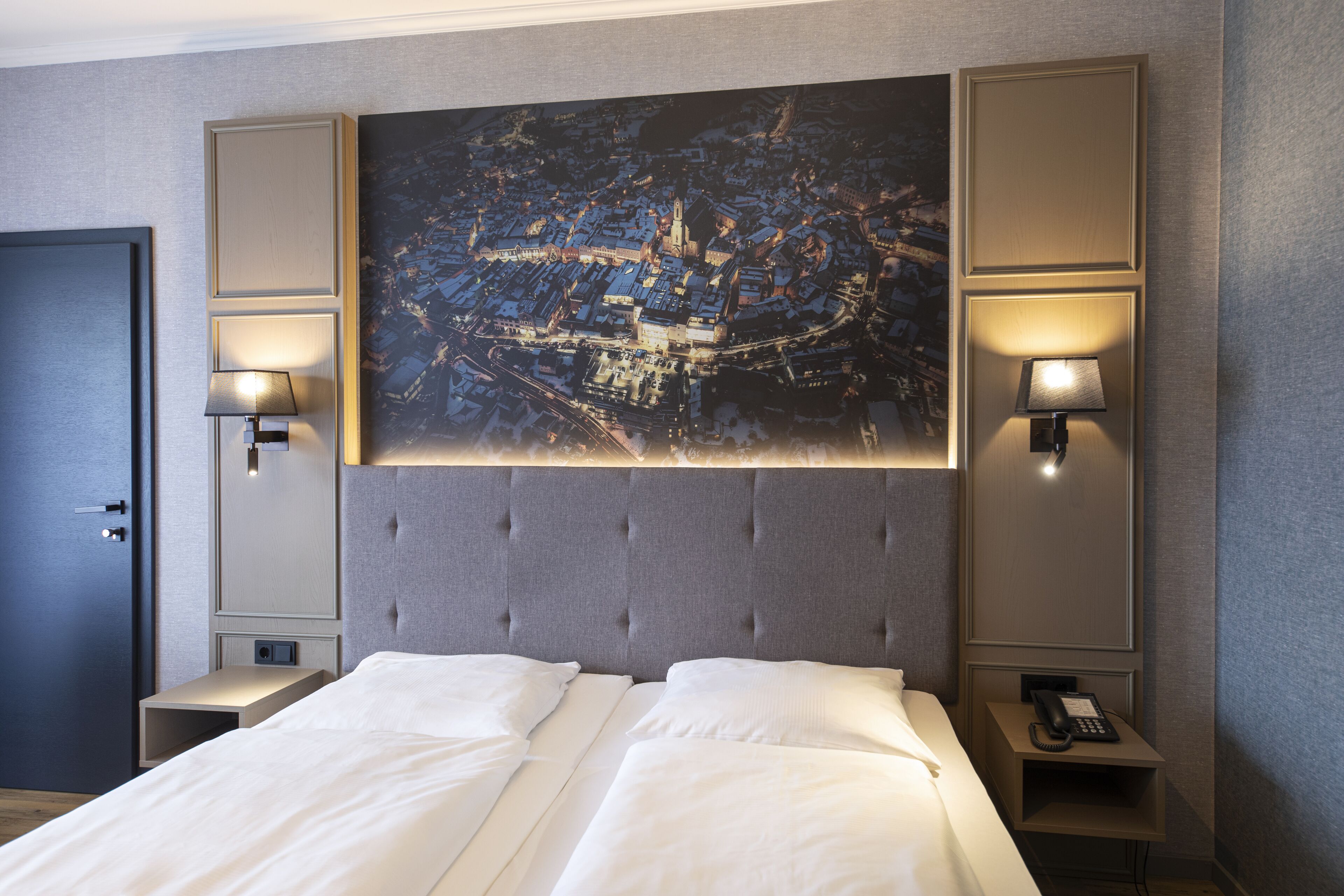 Una habitación de hotel con una cama grande, dos mesitas de noche, un cabecero con una pintura de un paisaje urbano y lámparas de pared.