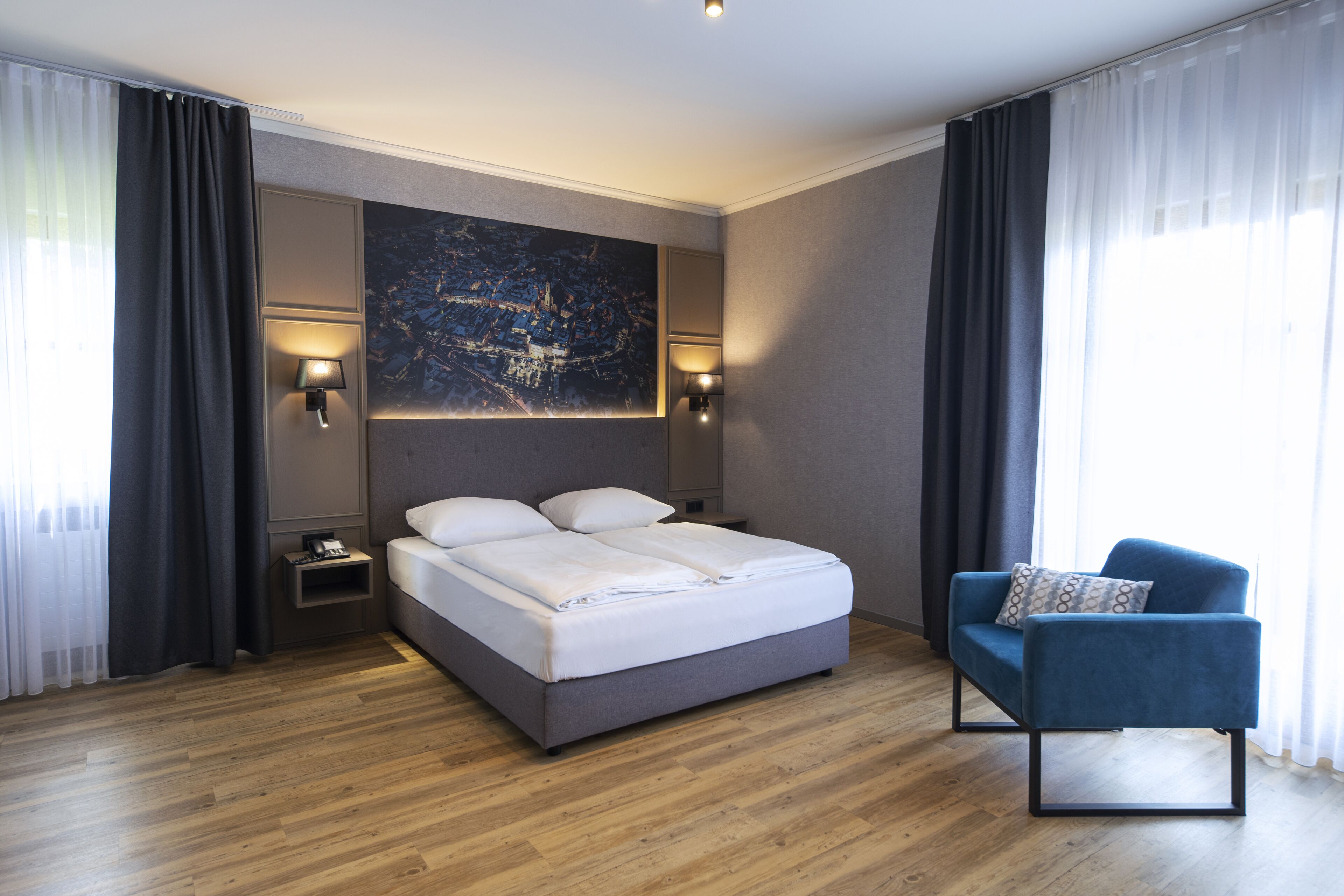 Ett modernt hotellrum med en stor säng, en blå fåtölj och en tavla med ett stadsmotiv på väggen.