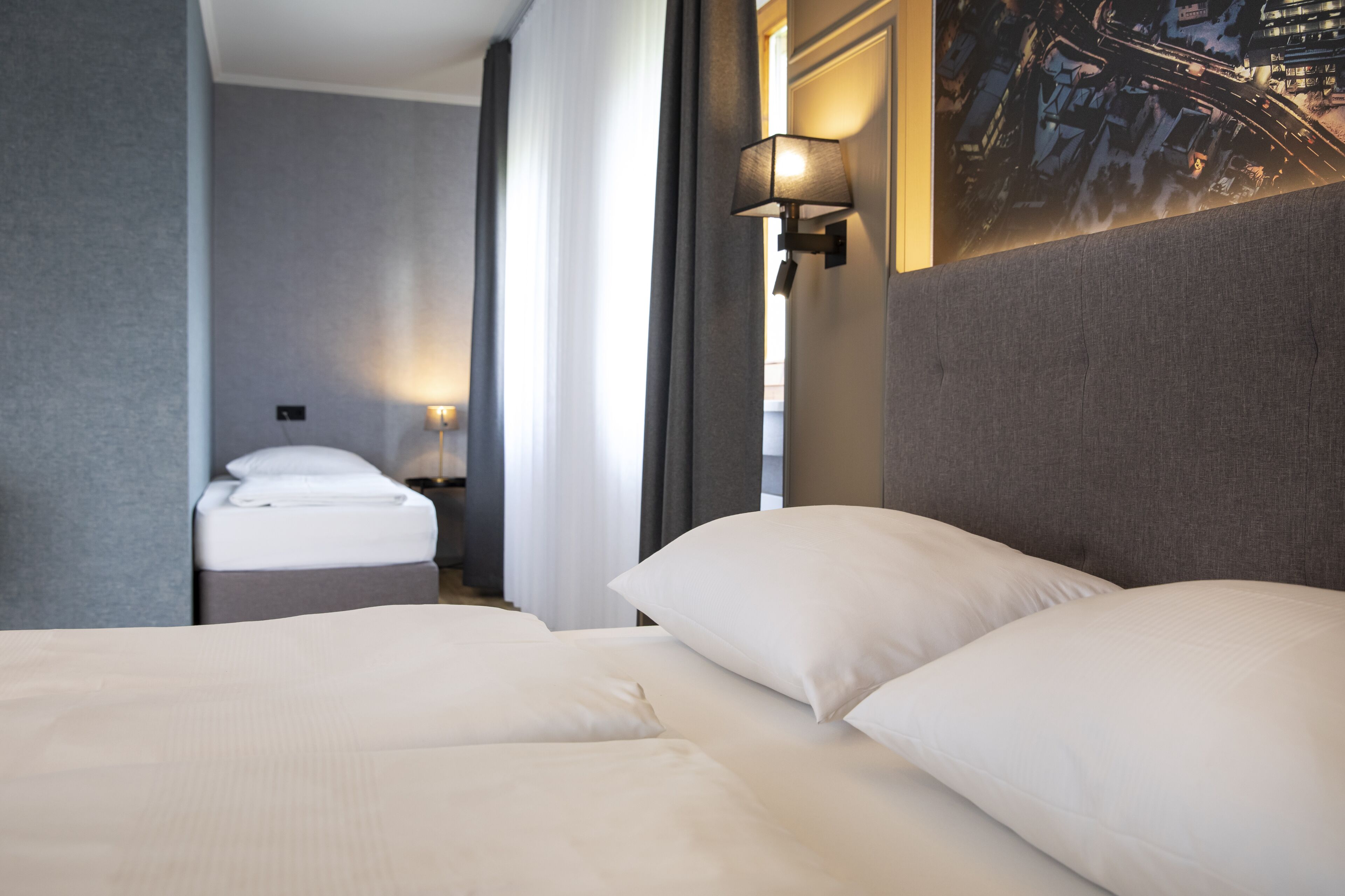 A hotel room with two beds, a nightstand, a lamp, and a cityscape painting on the wall.