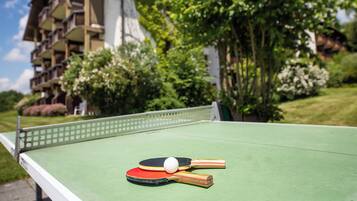Mesa de ping pong con dos paletas y una pelota frente a un edificio con balcones y vegetación.