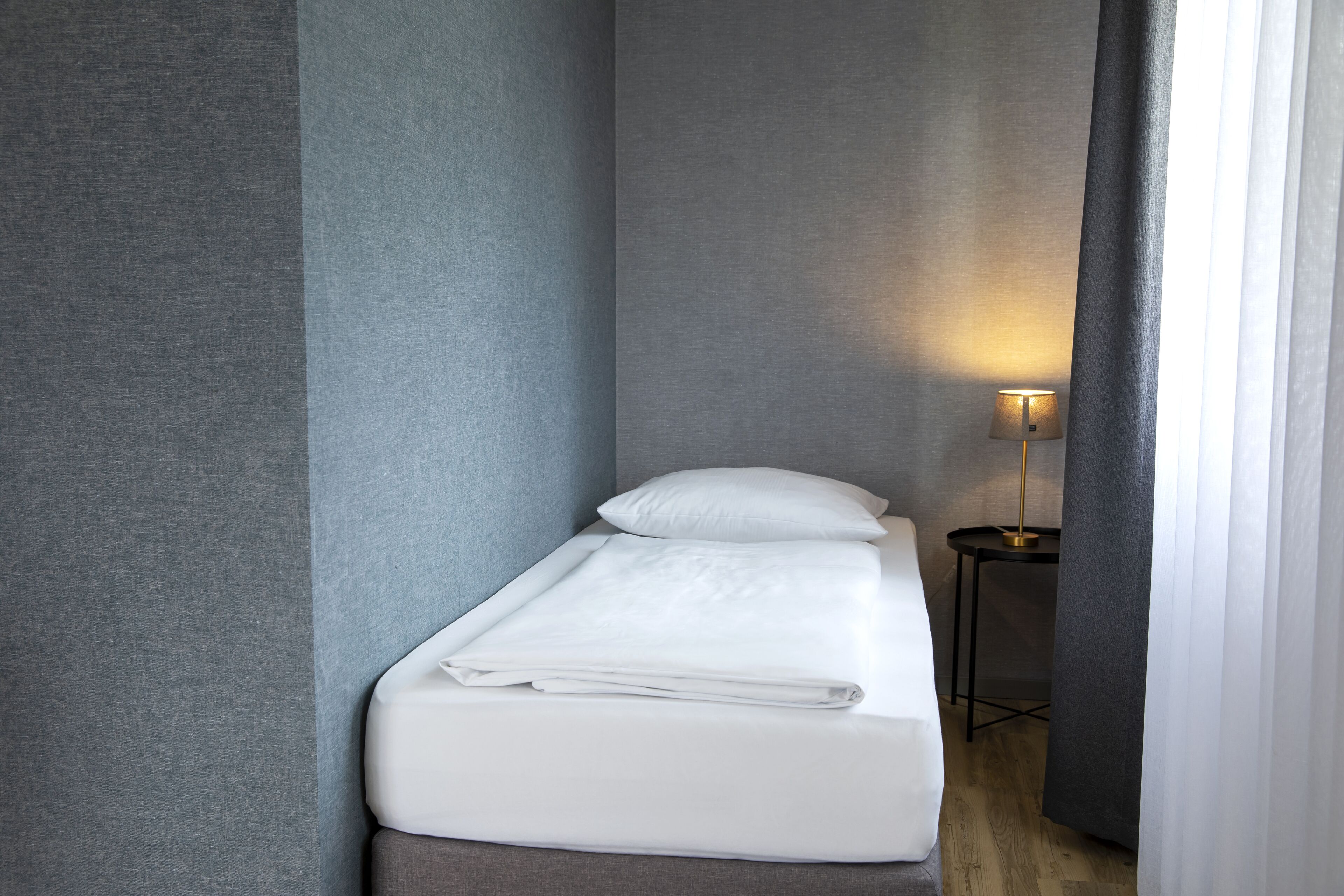 A small, neatly made bed with white linens in a compact room with grey walls and a small table with a lamp.