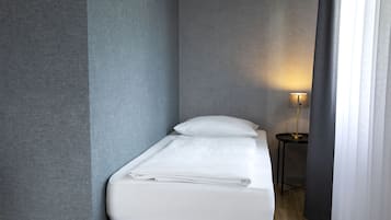 A small, neatly made bed with white linens in a compact room with grey walls and a small table with a lamp.
