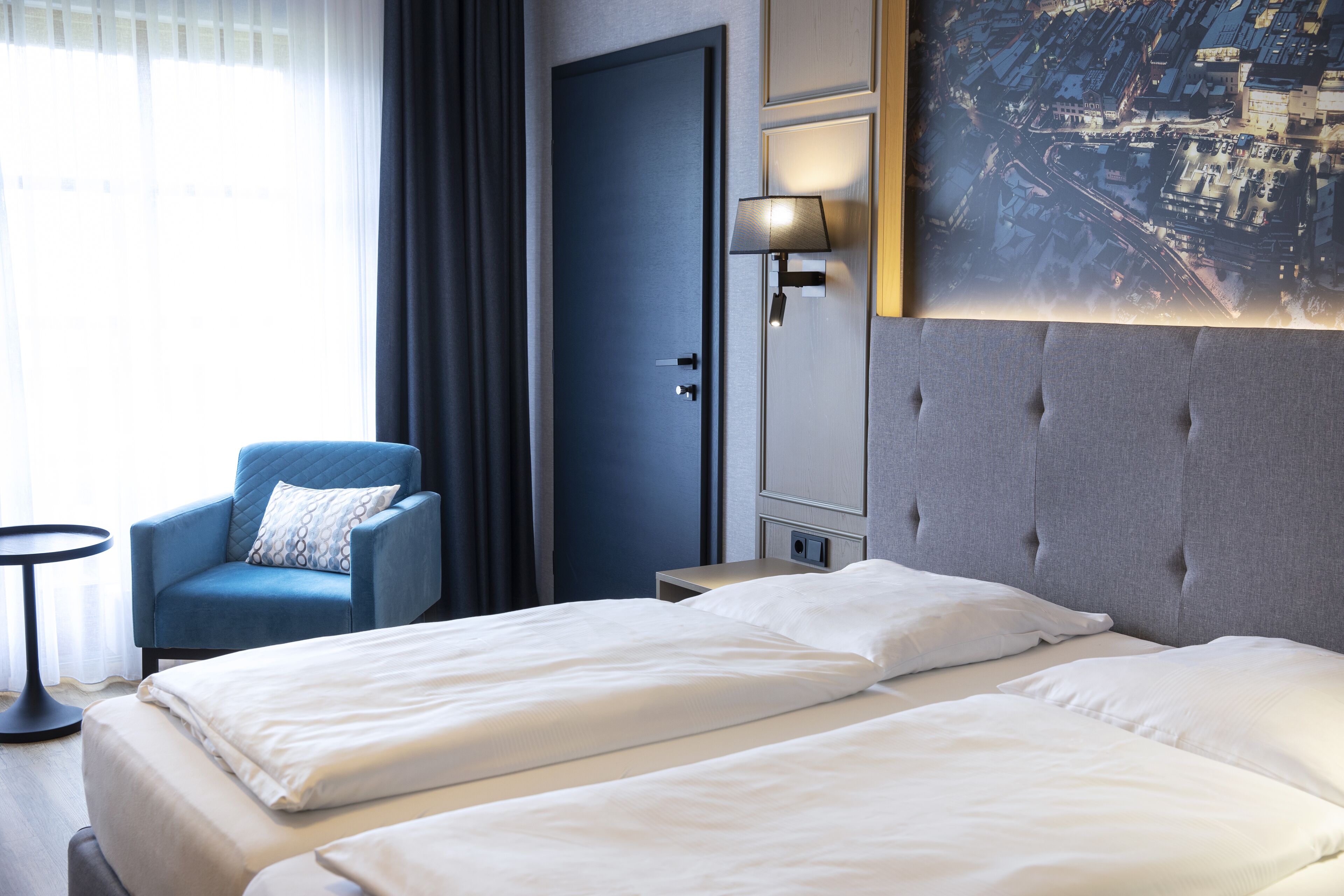 A hotel room with two beds, a blue armchair, a nightstand, a lamp, and a cityscape painting on the wall.
