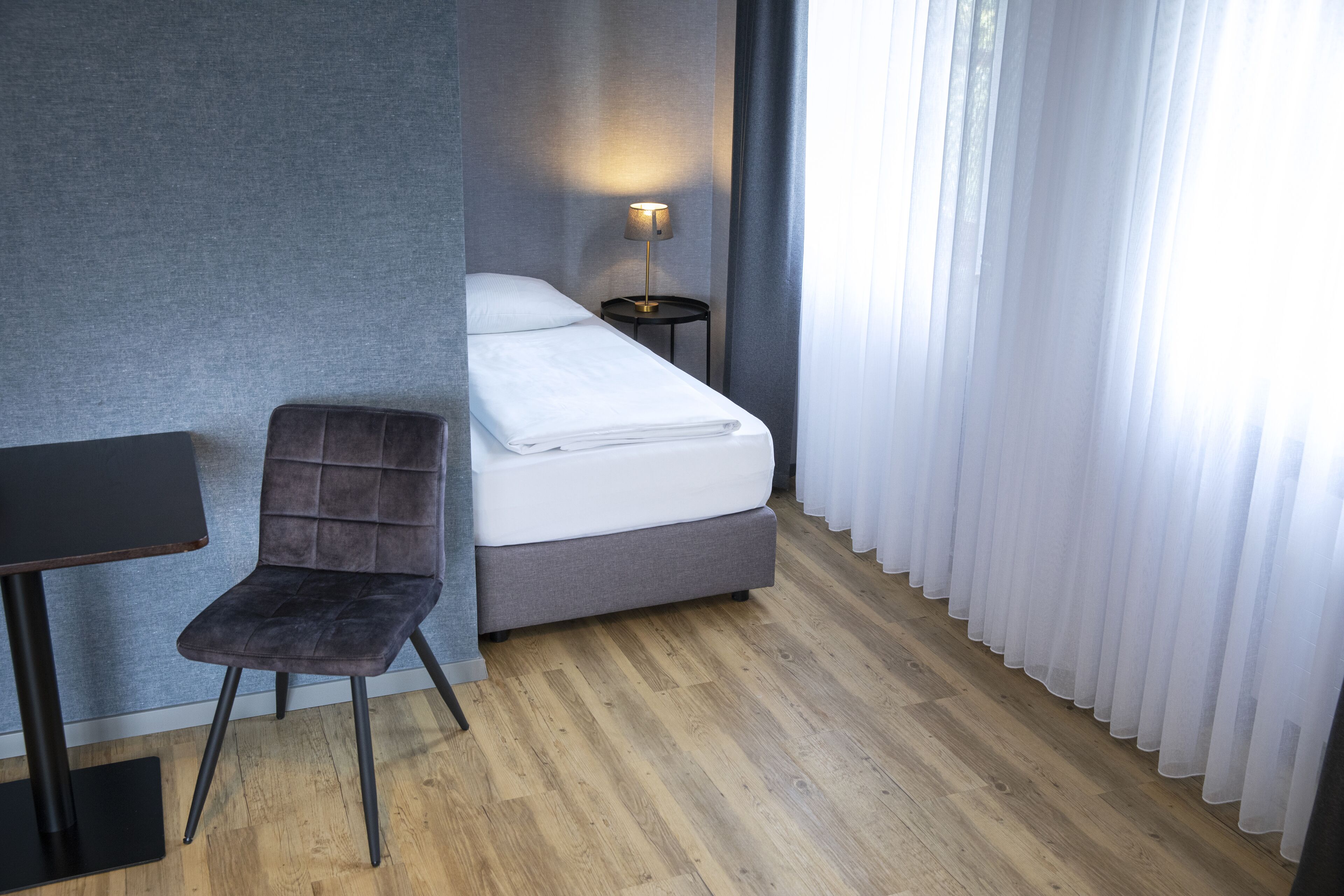 A hotel room with a bed, a chair, a small table, and a lamp.