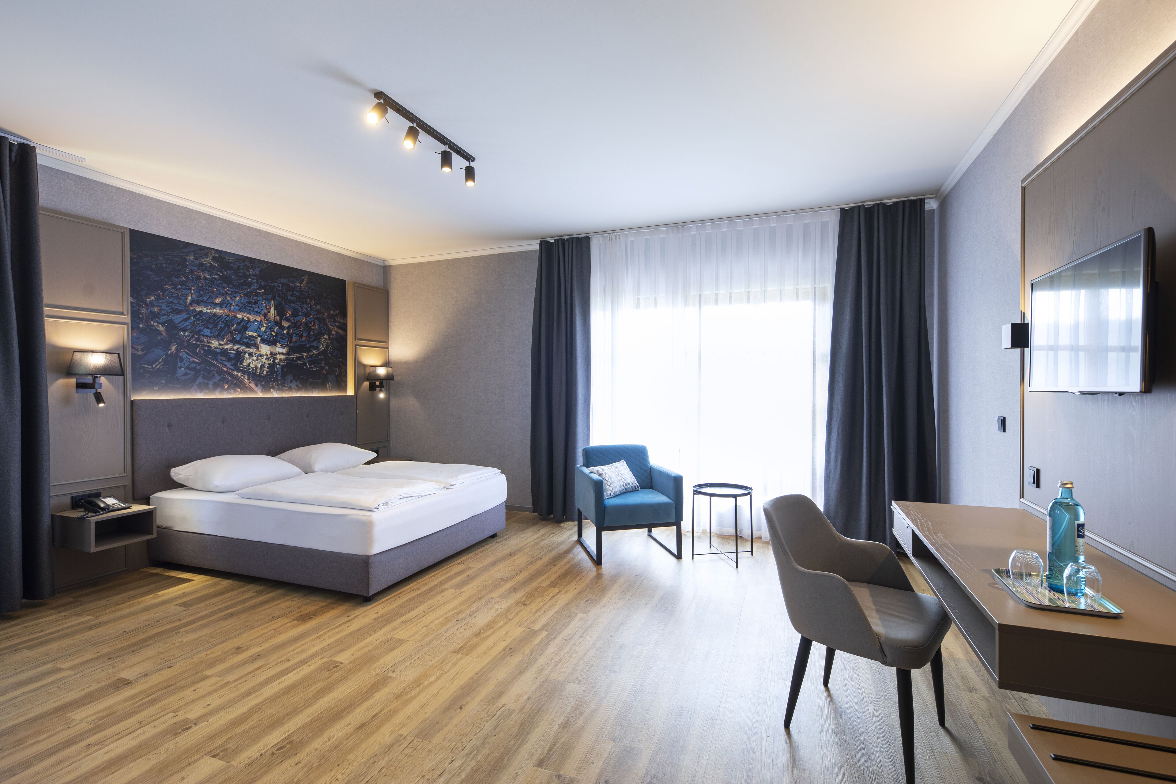 Une chambre d’hôtel moderne équipée d’un lit, d’un bureau, de deux chaises, d’une télévision et d’une grande fenêtre avec des rideaux.