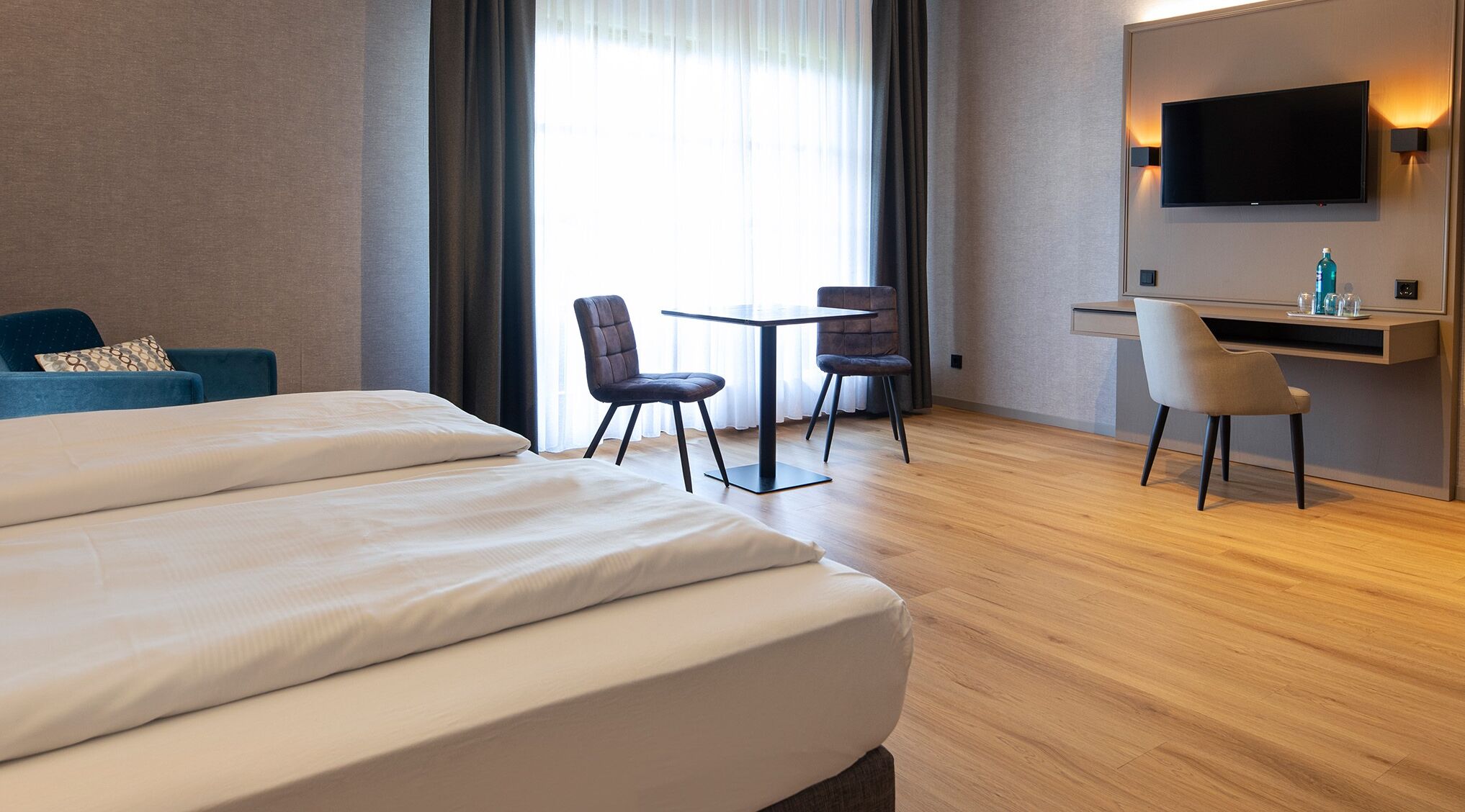Quarto de hotel com uma cama grande, duas cadeiras, uma mesa pequena, uma escrivaninha com um computador e uma TV fixada na parede.