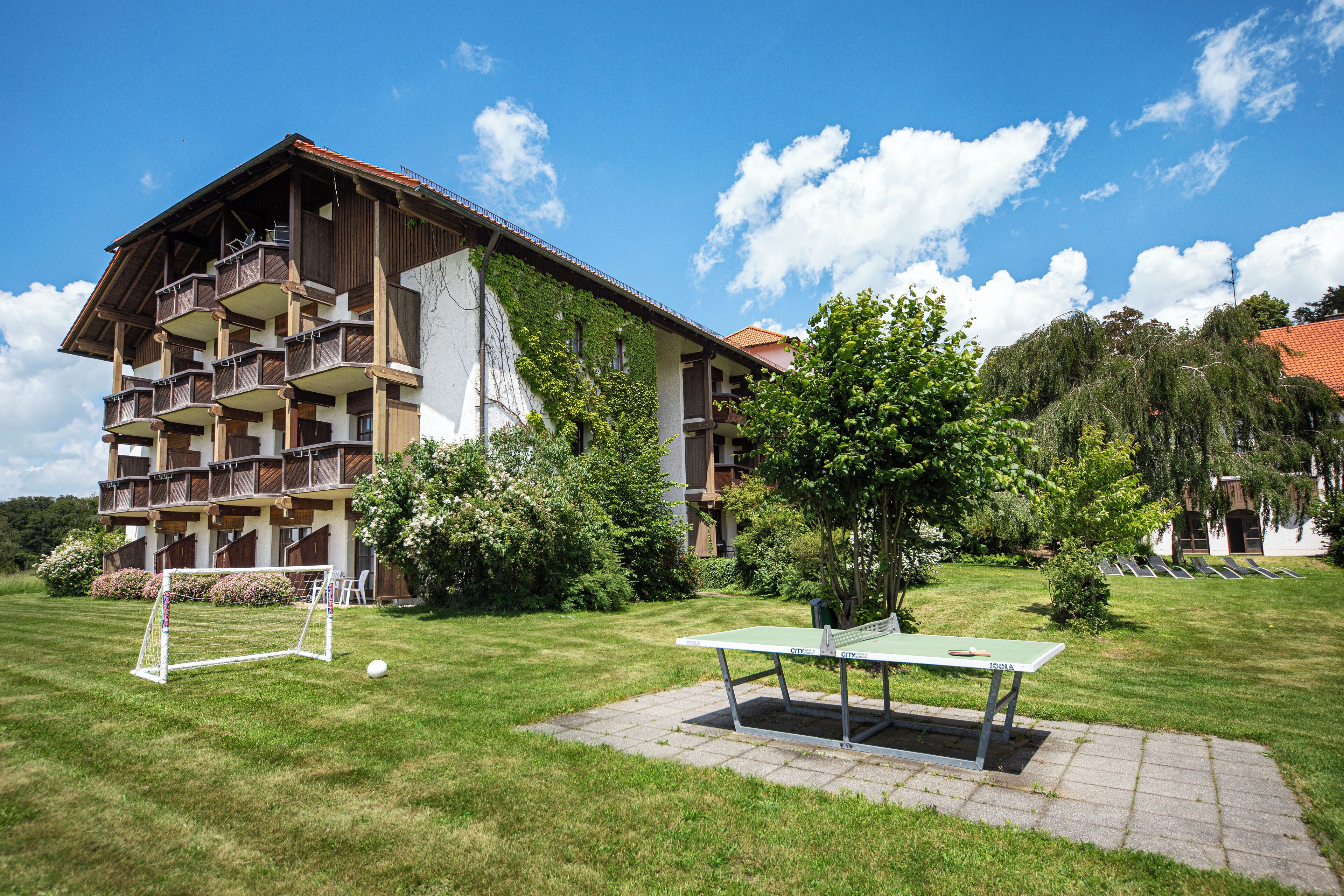 Un bâtiment doté de nombreux balcons, un espace verdoyant avec une table et un poteau de but, et un ciel bleu dégagé.