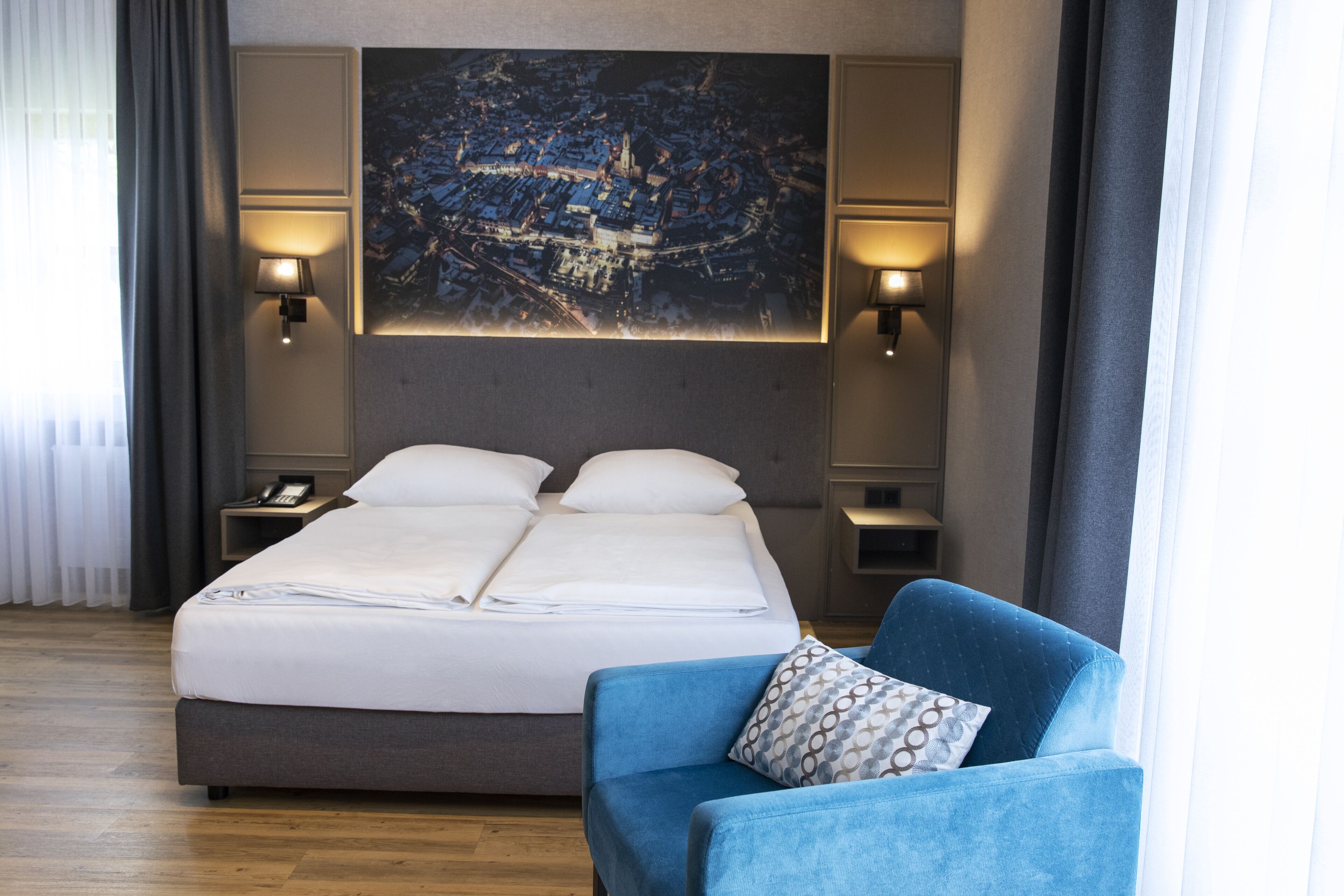 Una habitación de hotel con una cama doble, dos mesitas de noche, un sillón azul y un cuadro grande con una vista urbana.