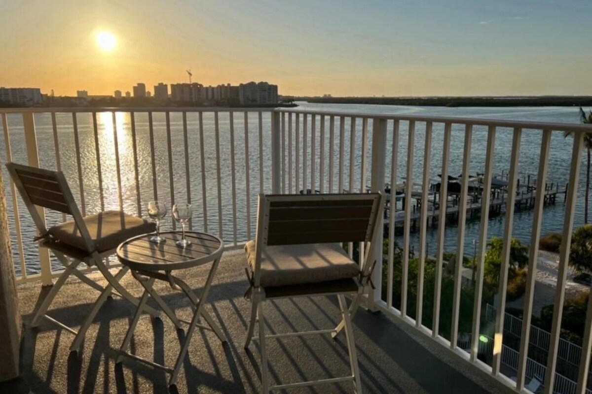 A balcony with two chairs and a small table, overlooking a body of water with a city skyline in the background.