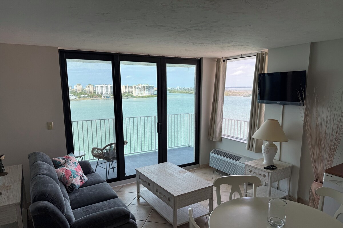 A living room with a sofa, a table, and a chair. There is a balcony with a view of the water and buildings.